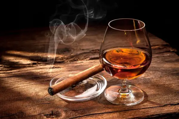 Ein Glas bernsteinfarbener Bourbon mit aufsteigendem Rauch von einer Zigarre auf einem Holztisch.