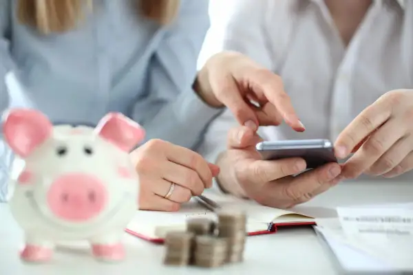 Dwie osoby planują wydatki, korzystając z telefonu i notatnika. Obok stoi skarbonka w kształcie świnki, symbolizująca oszczędności i fundusz socjalny.