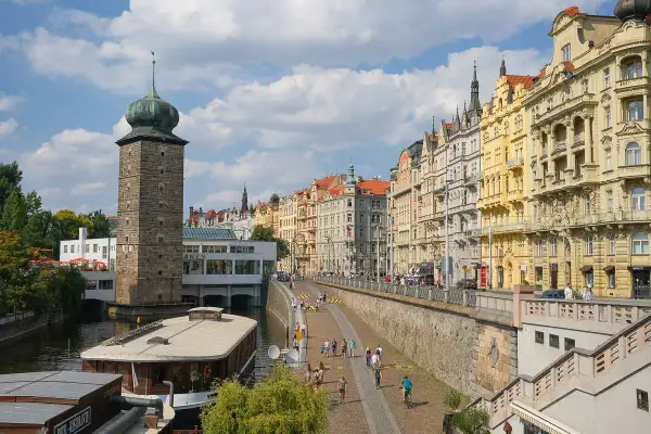 Vue de Prague que voir : tour de pierre avec dôme vert, maisons colorées le long de la rivière, et une péniche.