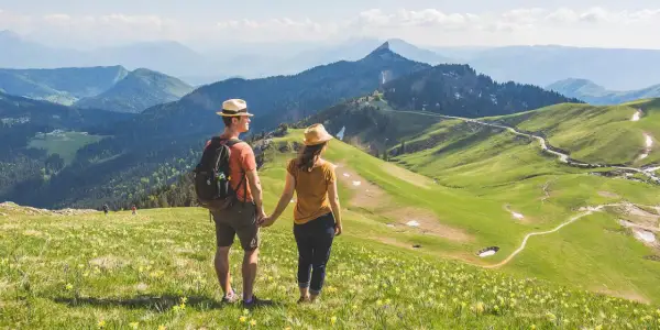 Couple en chapeau admirant un paysage de montagne verdoyant, parsemé de fleurs jaunes. Une rando Chartreuse insolite, avec des sentiers sinueux et des sommets bleutés à l'horizon.
