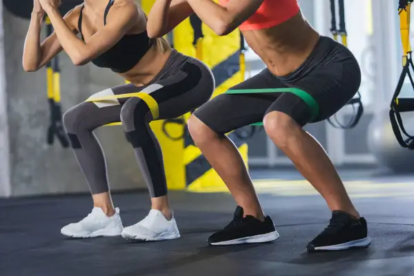Dwie kobiety wykonują ćwiczenia z gumami oporowymi na udach, przypominające przysiady. Nie jest to hack squat maszyna, ale ćwiczenie angażujące podobne partie mięśni.