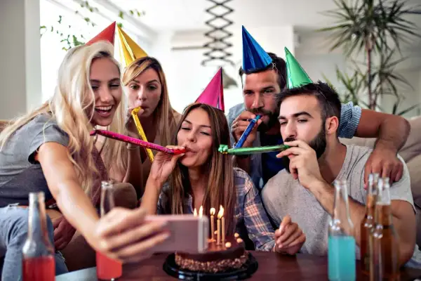 Grupa przyjaciół świętuje 70 urodziny, dmuchając w trąbki i robiąc selfie przy torcie.