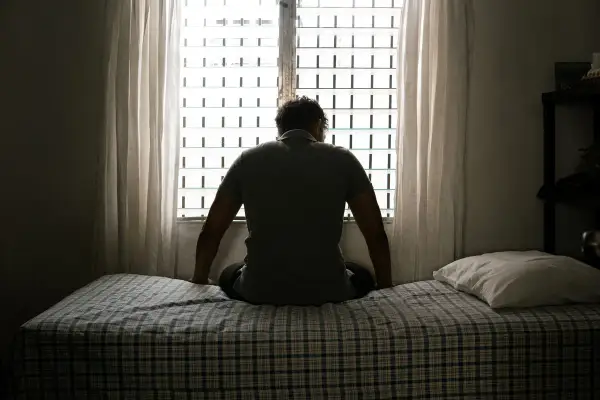 Hombre sentado en la cama, mirando por la ventana. Parece sumido en sus pensamientos, atravesando una **noche oscura del alma**.