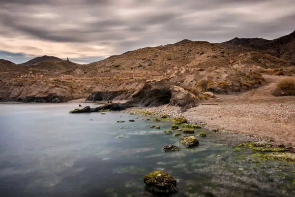 Mar tranquilo en la cala invencible Almería, con rocas cubiertas de algas y ruinas en la ladera.