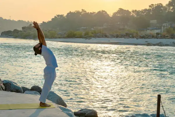 Hombre practica yoga Sivananda en la orilla de un río al amanecer, con los brazos extendidos hacia el cielo.