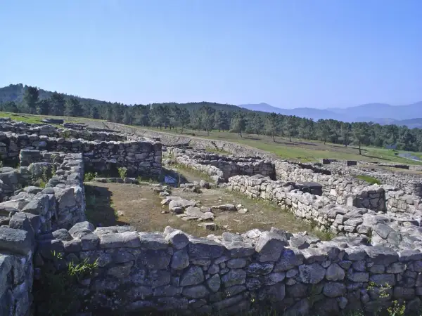 Ruinas del castro de San Cibrao de Las, con muros de piedra y un paisaje boscoso al fondo bajo un cielo azul.