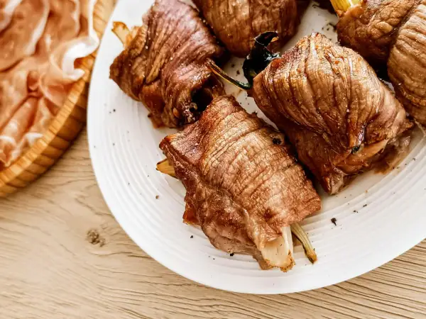 Involtini di carne succulenti, cotti alla perfezione, serviti su un piatto bianco. A lato, fette di prosciutto crudo in un cestino di legno.
