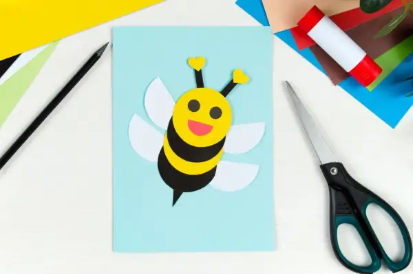 Ein süßes Biene basteln-Projekt auf hellblauem Papier. Bunte Bastelmaterialien, Schere und Klebestift liegen bereit.