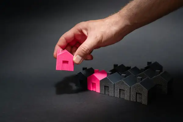 Mano che sceglie una casetta rosa tra tante nere. Un'idea di permuta immobiliare con costruttore.