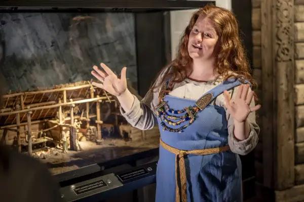 En kvinna i vikingakläder berättar vid en modell av en vikingagård på ett museum i Stockholm.