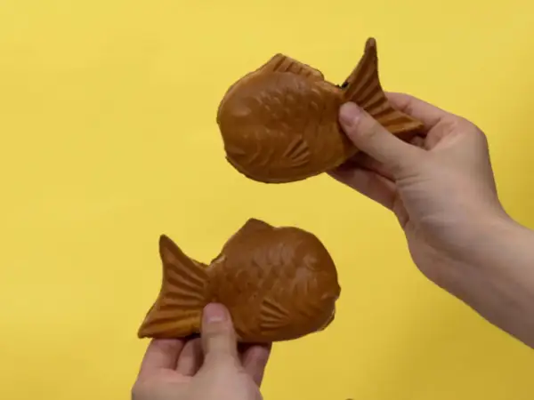 Zwei goldbraune Taiyaki, beliebte japanische Desserts in Fischform, werden von Händen auf gelbem Hintergrund gehalten.