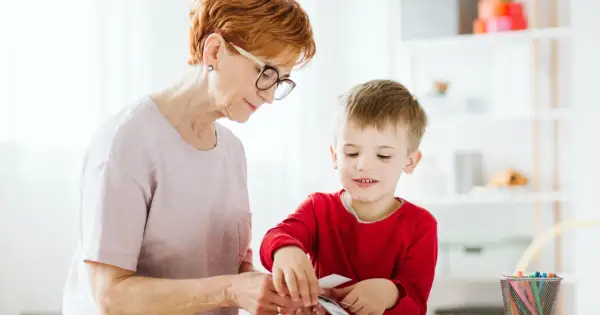 Babcia i wnuczek tworzą coś razem. Dziecko z zaciekawieniem przygląda się swoim ruchom, a jego manieryzmy ruchowe są widoczne w skupieniu.