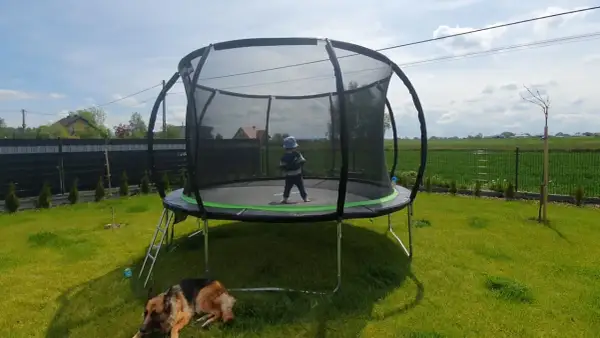 Dziecko w czapce na trampolinie, pies odpoczywa na trawie. Czas na wesołe zabawy na trampolinie w ogrodzie.