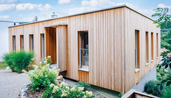 Modernes Haus mit einer Mischfassade aus Holz und Putz. Vertikale Holzverkleidung umrahmt schmale Fenster. Grüne Pflanzen schmücken den Eingangsbereich.