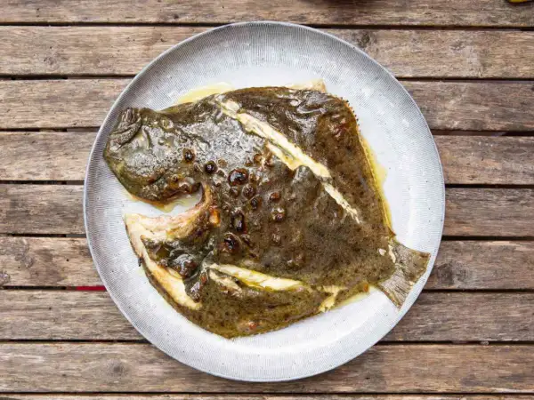 A whole grilled turbot, glistening with butter and herbs, served on a rustic plate atop a weathered wooden table.