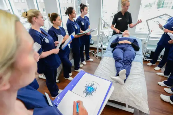 Elever på hudterapeut utbildning distans lyssnar på instruktör. En elev ligger på en brits medan andra antecknar.