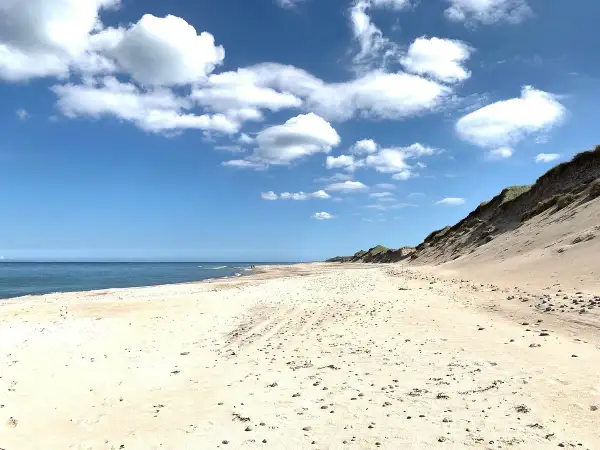 Sandy beach with dunes, calm blue sea, and white clouds under a bright blue sky.