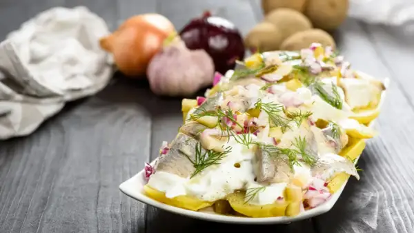 Pyszna sałatka ziemniaczana ze śledziem, udekorowana koperkiem i czerwoną cebulką. Obok leżą ziemniaki, cebula i czosnek.
