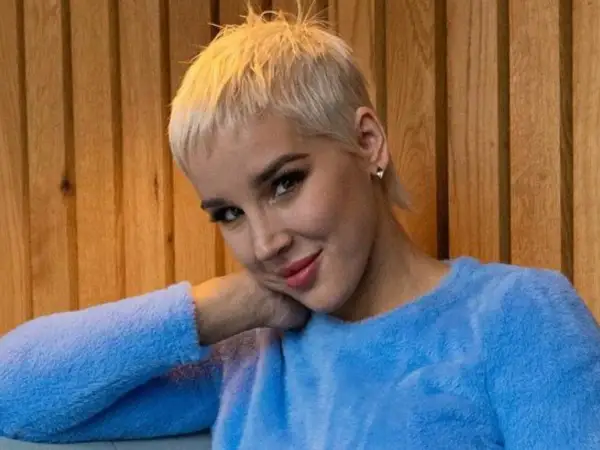 Kobieta z krótkimi blond włosami w niebieskim swetrze opiera głowę na dłoni.