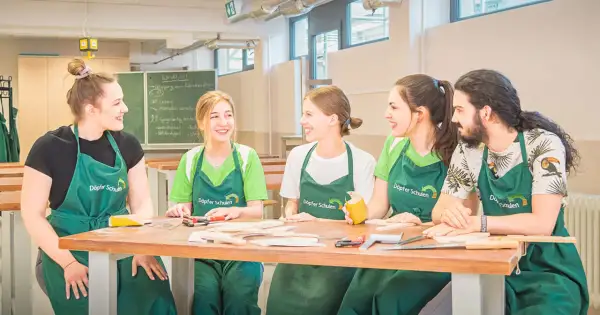 Fünf junge Menschen in grünen Schürzen lachen und arbeiten an einem Holztisch. Hier beginnt ihre kostenfreie Ausbildung Ergotherapie.