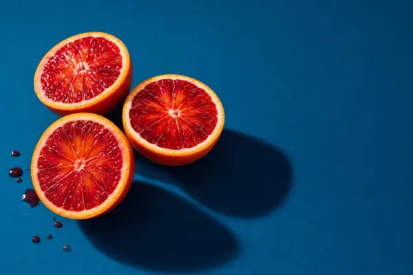 Vibrant fruit photography: three blood oranges halved, revealing their deep red flesh, with a few juice droplets on a deep blue background.
