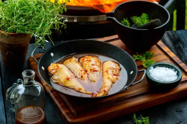 Kycklingvingar i en glansig, kryddig egen marinad i en stekpanna, redo för grillen. Bredvid finns färska örter och flingsalt.