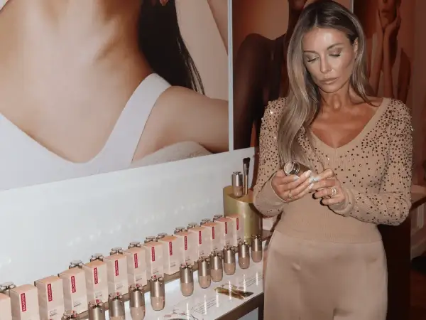 Kobieta z długimi blond włosami trzyma buteleczkę Clarins Double Serum Foundation. Na półce stoją dziesiątki opakowań tego podkładu.
