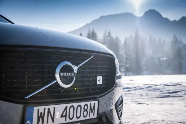 Szary Volvo XC60 Polestar z emblematem Polestar na grillu, zaparkowany na tle ośnieżonych gór i lasu.
