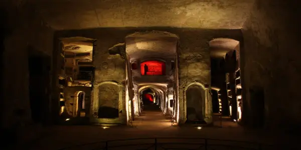 Corridoio sotterraneo illuminato, con nicchie scavate nella roccia e un'apertura rossa in lontananza, evocando l'atmosfera della tomba di San Gennaro.