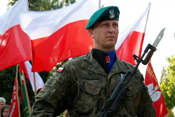 Dumnie stoi dowódca w zielonym beretcie, z karabinem i polską flagą w tle.