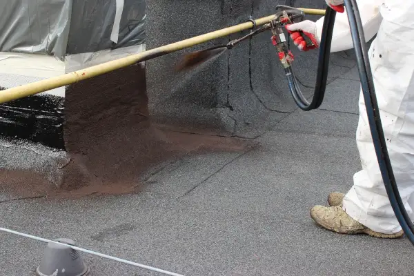 Pracownik w białym kombinezonie natryskuje ciemną masę na dach, uszczelniając go. To praca z **styropapą**.