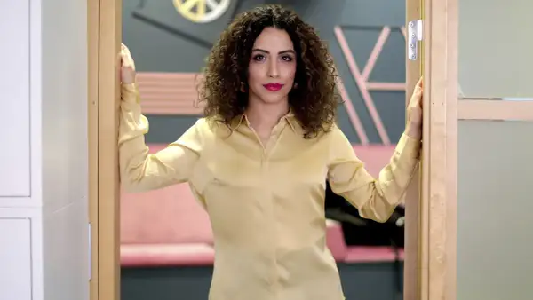 Helya Riazat, med lockigt mörkt hår och rött läppstift, står i en dörröppning i en guldig blus.