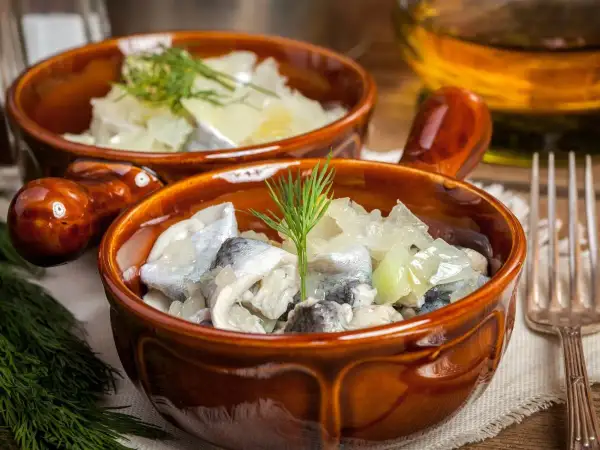 Dwa brązowe naczynia z pysznymi śledziami na słodko, udekorowane gałązkami koperku. Obok widelczyk i butelka z oliwą.