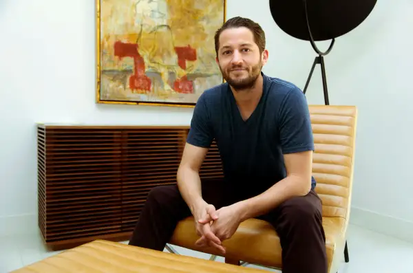 Ian Kirkpatrick, a man with a beard and a blue t-shirt, sits in a tan leather chair in a modern room with abstract art.
