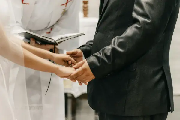 Mani che si stringono durante la dichiarazione matrimonio, con un libro aperto e un sacerdote sullo sfondo.
