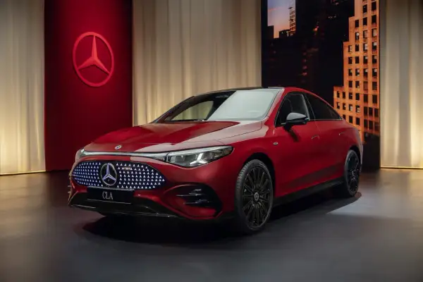 Elegancki, czerwony nowy Mercedes CLA z podświetlanym grillem i czarnymi felgami stoi na ciemnym tle z miejskim krajobrazem w tle.