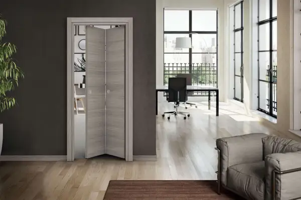 Montaggio porte interne a soffietto in legno chiaro, che dividono un soggiorno moderno da un ufficio luminoso con scrivania e poltrona.