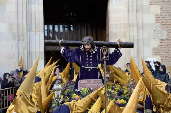 Imagen de una **procesión Miércoles Santo**. Una figura de Jesús carga la cruz, rodeada de nazarenos con túnicas moradas y capirotes dorados.