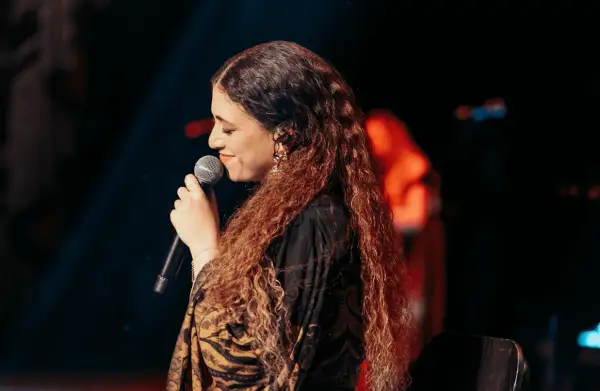 Cantante con lunghi capelli ricci, microfono in mano, sul palco. Un'espressione che evoca la bellezza della Sicilia, un testo che risuona.