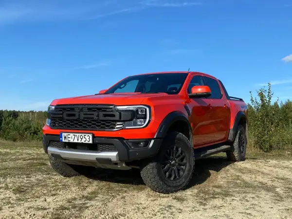 Pomarańczowy Ford Ranger Raptor gotowy na przygodę na piaszczystym terenie, z lasem w tle i błękitnym niebem.