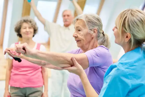 Seniorzy wykonują ćwiczenia na alzheimera z fizjoterapeutką. W tle inni uczestnicy ćwiczą z hantlami.