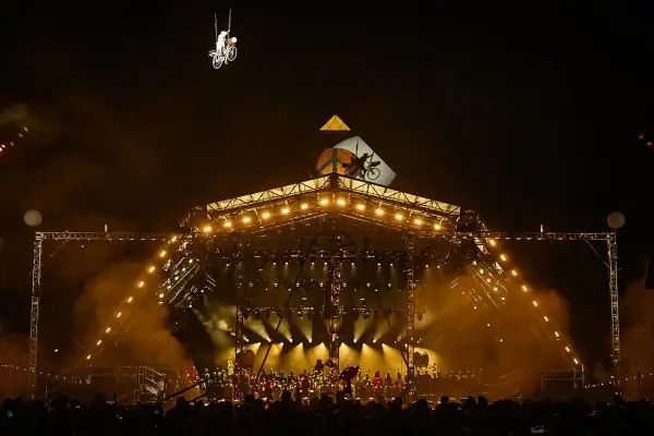 A performer on a bicycle is suspended in the air above a brightly lit stage at Glastonbury. The crowd below anticipates the next act, checking their Glastonbury set times.