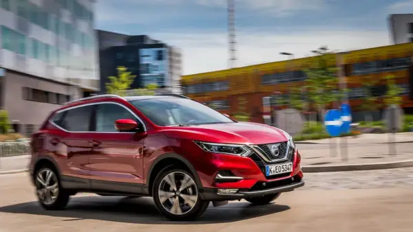 Czerwony Nissan Qashqai 1 jedzie ulicą w mieście. W tle nowoczesne budynki i niebieski znak drogowy.
