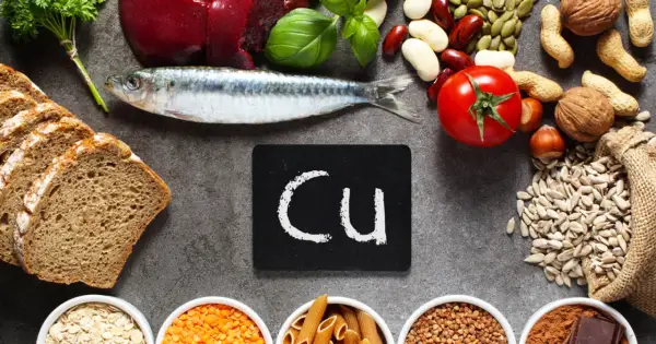Na stole leżą ryba, chleb, pomidor, orzechy, nasiona i inne produkty bogate w miedź. Tabliczka z napisem "Cu" symbolizuje ten pierwiastek.