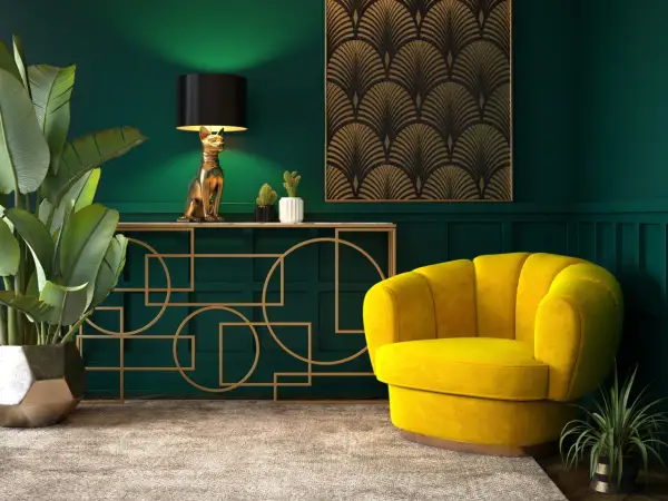 Elegancka tkanina art deco zdobi ścianę nad złotą konsolą z lampą w kształcie kota. Obok żółty fotel i bujna roślina.
