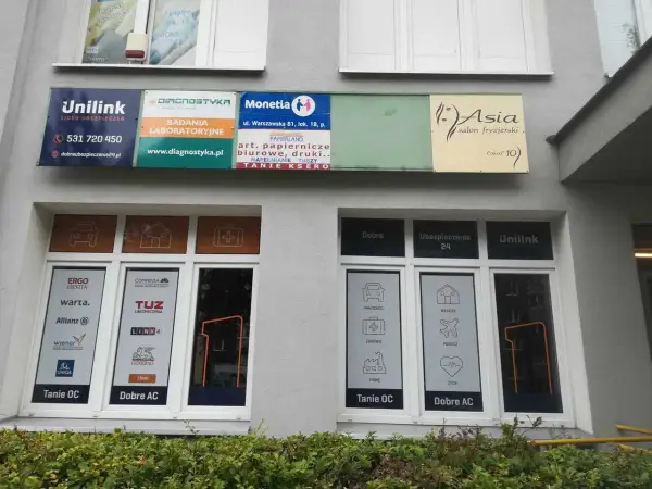 Szyldy firm ubezpieczeniowych i salonu fryzjerskiego na szarej ścianie budynku.
