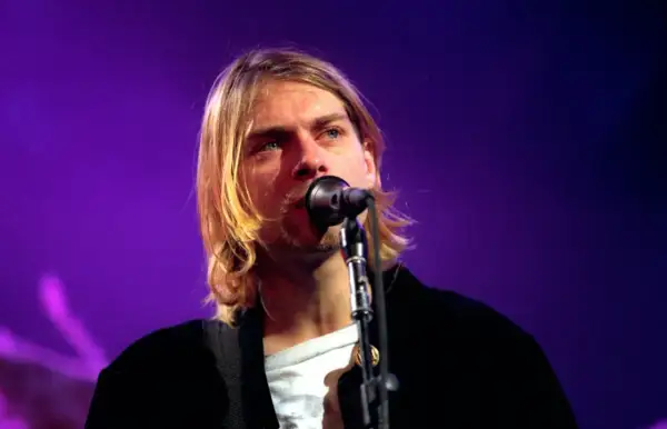 Kurt Cobain śpiewa do mikrofonu na tle fioletowej sceny.