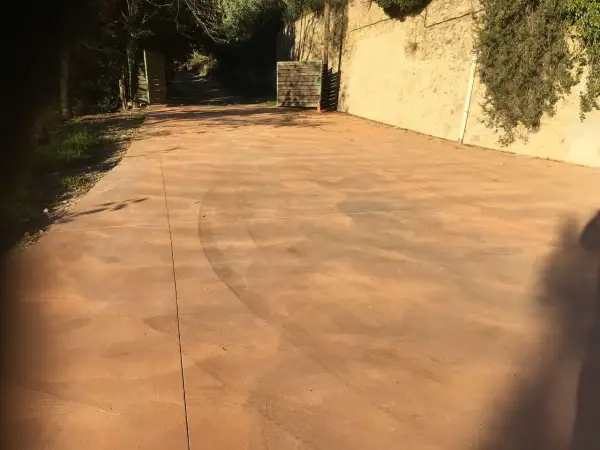 Un camino de hormigón de colores, con marcas de neumáticos, se extiende hacia una entrada flanqueada por vegetación y un muro de piedra.