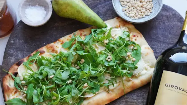 En läcker pinsa med chevre, ruccola och pinjenötter, bredvid en päronhalva, salt och en vinflaska.