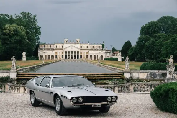Srebrny Lamborghini Espada przed okazałą willą z basenem i rzeźbami. Klasyk motoryzacji w malowniczej scenerii.